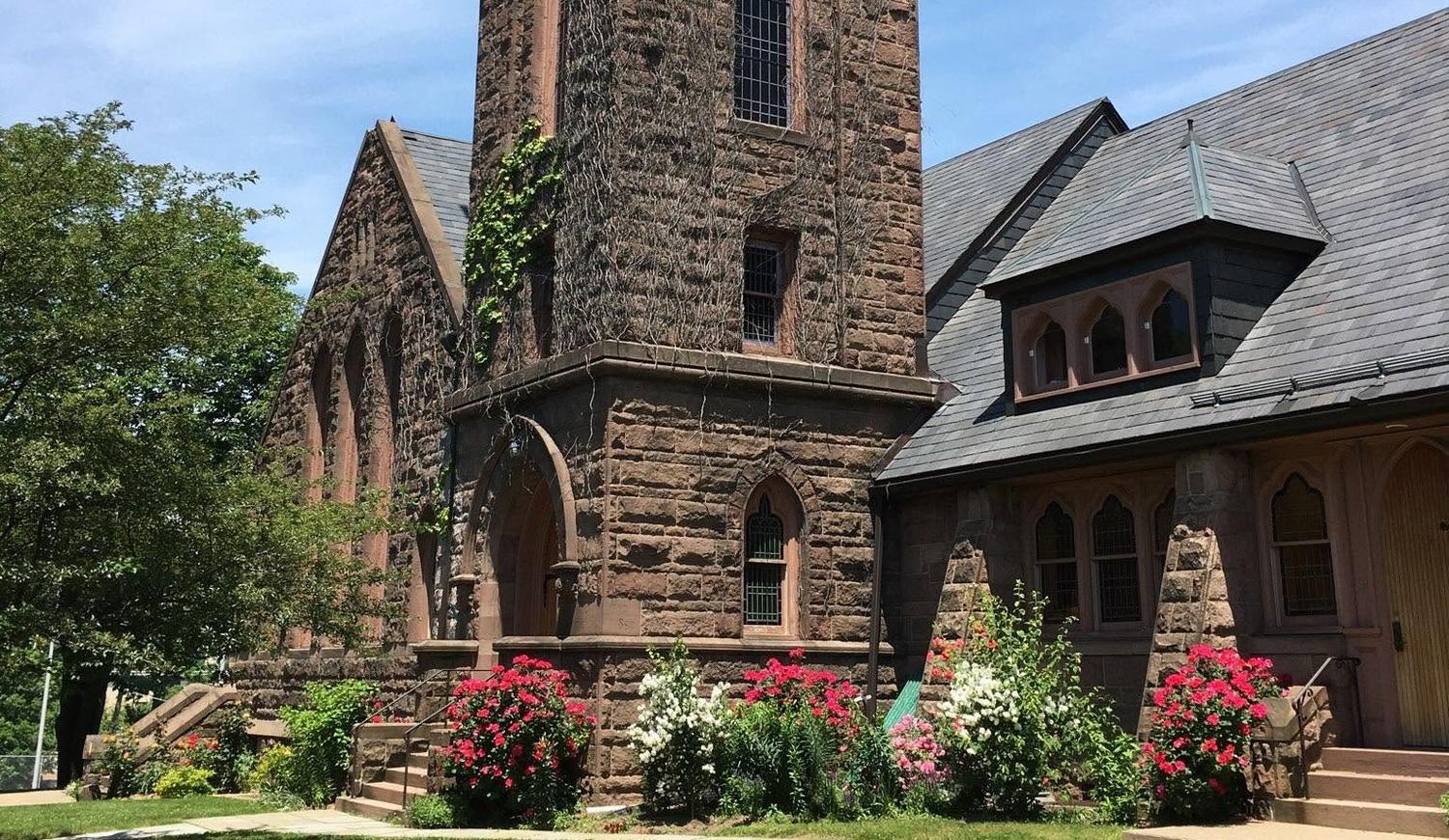 The brownstone tower and slate roofline of First Baptist Church of Morristown on a summer afternoon, with the front garden in bloom.